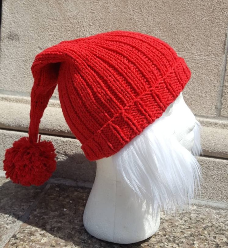 Puede incluir: Un gorro rojo de punto con una larga punta puntiaguda y un pomp&oacute;n en el extremo. El gorro est&aacute; en una cabeza de maniqu&iacute; blanca con una barba blanca.