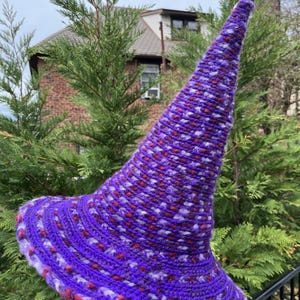 May include: A handmade, purple crochet witch hat with a wide brim and a tall, pointed crown. The hat features a textured pattern with red and white flecks throughout. The brim is trimmed with a lighter purple yarn.
