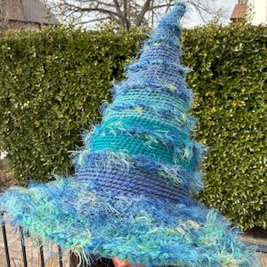 May include: A handmade witch hat in shades of blue and teal. The hat features a wide brim and a tall, pointed crown. The hat is made of a textured yarn with a fuzzy, fringed trim.