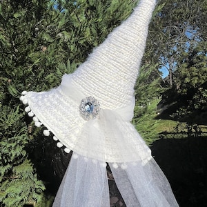 May include: A white witch hat with a wide brim, adorned with a sheer veil, a ribbon, and a sparkling jewel. The hat features a textured knit design and small white pom-poms along the brim, creating a whimsical and elegant look.