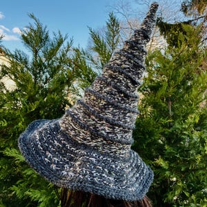 May include: A hand-knitted witch hat with a tall, pointed crown and a wide brim. The hat is made of a textured yarn in shades of gray, white, and flecks of other colors. The hat is a unique accessory for Halloween or costume events.