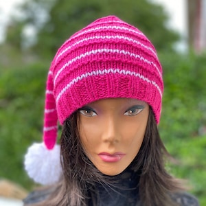 May include: A knitted hat in a vibrant fuchsia color with horizontal white stripes. The hat has a long, pointed tip and a white pom-pom. The hat is designed to be worn on the head.