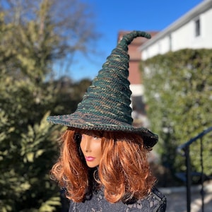 May include: A green and brown crocheted witch hat with a tall, pointed crown and a wide brim. The hat is worn by a mannequin with long, red hair.