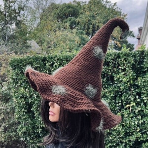 May include: A brown crocheted witch hat with a pointed crown and a wide brim. The hat has green fuzzy accents on the brim and crown.