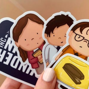 May include: Three cartoon stickers featuring characters from the TV show "The Office". The stickers are on a white background. The first sticker is of a woman with brown hair wearing a red shirt and holding a coffee cup. The second sticker is of a man with brown hair wearing a white shirt and tie. The third sticker is of a man with brown hair wearing an orange shirt and glasses. He is holding a yellow gelatin mold with a brown stapler inside. The text "Dunder Mifflin Inc." is visible on the bottom left corner of the image.