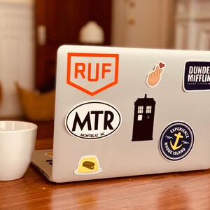 May include: A silver laptop computer with various stickers on the lid. The stickers include a red and orange logo with the letters "RUF", a black and white oval sticker with the text "MTR MONTREAT NC", a black silhouette of a TARDIS, a blue and yellow sticker with an anchor and the text "EXPERIENCE RHODE ISLAND", a pink hand with a heart, and a blue sticker with the text "DUNDER MIFFLIN PAPER COMPANY".