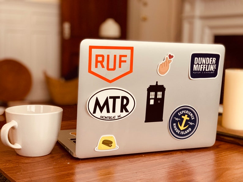 May include: A silver laptop computer with various stickers on the lid. The stickers include a red and orange logo with the letters "RUF", a black and white oval sticker with the text "MTR MONTREAT NC", a black silhouette of a TARDIS, a blue and yellow sticker with an anchor and the text "EXPERIENCE RHODE ISLAND", a pink hand with a heart, and a blue sticker with the text "DUNDER MIFFLIN PAPER COMPANY".