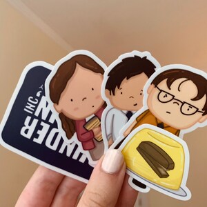 May include: Three cartoon stickers featuring characters from the TV show "The Office". The stickers are on a white background. The first sticker is a blue and white sticker with the text "UNDER IN INC." The second sticker is a cartoon of a woman with brown hair wearing a red shirt and holding a stack of papers. The third sticker is a cartoon of a man with brown hair wearing a yellow shirt and holding a plate of yellow jello with a brown chocolate bar inside.