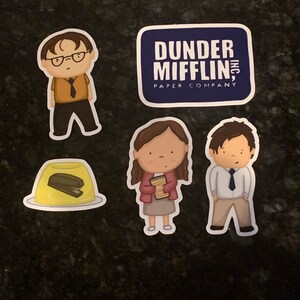 May include: Four stickers featuring characters and a logo from the TV show "The Office". The stickers are on a dark gray surface. The stickers feature a character with glasses, a character holding a stack of papers, a character with a tie, and a logo that says "Dunder Mifflin, Inc. Paper Company".
