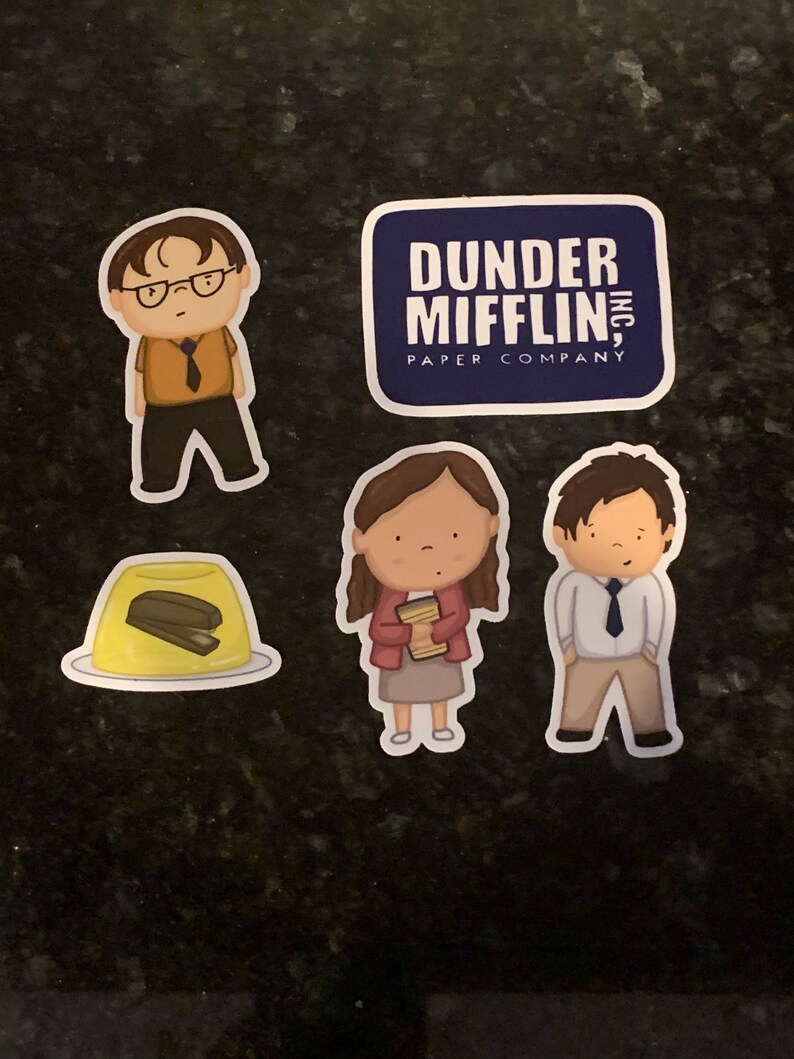 May include: Four stickers featuring characters and a logo from the TV show "The Office". The stickers are on a dark gray surface. The stickers feature a character with glasses, a character holding a stack of papers, a character with a tie, and a logo that says "Dunder Mifflin, Inc. Paper Company".