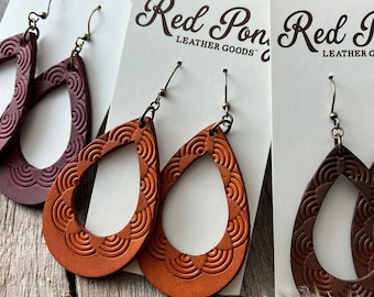 May include: Three pairs of leather earrings with a scalloped design. The earrings are displayed on white cards with the text "Red Pony Leather Goods". The earrings are in brown, burgundy, and orange.