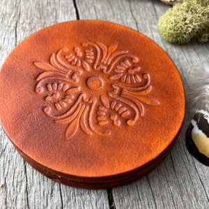 May include: A stack of round, brown leather coasters. Each coaster features an embossed floral design in the center. The coasters have a rustic, vintage aesthetic, with variations in the leather's color and texture.