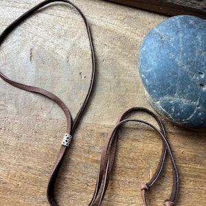Peut inclure: Un collier en cuir marron avec un accent argenté et des liens réglables. Le collier est présenté sur une surface en bois, avec une pierre bleu-gris en arrière-plan.