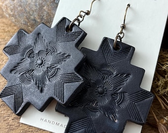 May include: Black leather earrings with a geometric design and a floral pattern. The earrings are handmade and feature a hook closure. The earrings are labeled "HANDMADE" and "S, TN".