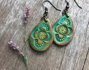 May include: A pair of teal and bronze teardrop earrings with an embossed floral design. The earrings have a rustic, antique appearance and are suspended from bronze-colored ear wires. A sprig of purple flowers is in the background.