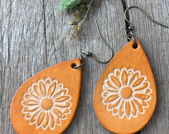 May include: A pair of orange leather teardrop earrings. Each earring has a daisy design embossed in the centre. The earrings have silver-coloured ear wires and are displayed on a wooden surface.