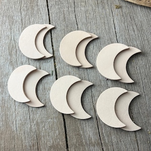 May include: Six unfinished wooden crescent moon shapes, each with a smaller crescent moon shape layered on top. The moons are arranged in a staggered pattern on a wooden surface.