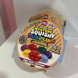 May include: A round cardboard packaging for a Mystery Squishy Dumpling toy. The label features the text "The Original Viral Mystery Squishy Dumpling" and "Rare Ruby Edition!" with illustrations of colorful, smiling dumpling characters.