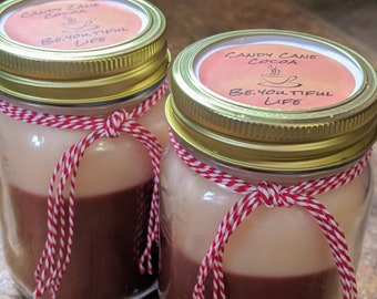 May include: Two glass jars with gold lids and red and white twine tied around them. The jars contain a layered candle with a white top layer and a brown bottom layer. The labels on the jars read "Candy Cane Cocoa" and "Be. You. Tiful Life".