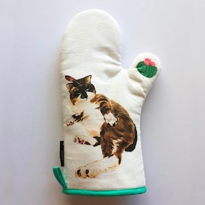 May include: White oven mitt with a green border. The mitt features a brown and white cat with a cactus on the top. The cat is looking to the left.