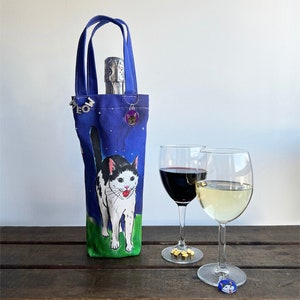 May include: A blue canvas wine bottle carrier with a black and white cat illustration. The cat is looking up with its mouth open. The carrier has a silver charm with a cat silhouette on it.