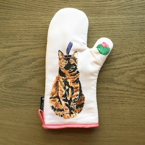 Puede incluir: Manopla de horno blanca con ribete rosa y diseño de gato. El gato es marrón, negro y naranja, con una pluma y una diadema. Un cactus verde con una flor rosa está en el pulgar.