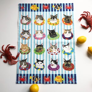 May include: A blue and white striped tea towel with a repeating pattern of cats wearing colorful life preservers. The cats are in various poses and have different colored fur. The towel has a border of blue and white stripes with images of starfish, crabs, fish, and seashells.