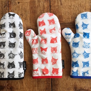 May include: Three oven mitts with cat face patterns. One mitt is white with black cat faces, another is white with red cat faces, and the third is white with blue cat faces. Each mitt has a solid-colored trim and a small black tag.
