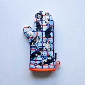 Op de afbeelding: Een ovenwant met een blauw-wit ruitpatroon met verschillende kattenillustraties in zwart, wit en grijs. De want heeft een oranje rand en een lus om op te hangen.