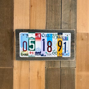 May include: A framed wall art piece made from repurposed license plates. The artwork displays the date "05 18 91" using license plates with various colors and designs. The frame is made of weathered wood, adding a rustic touch.