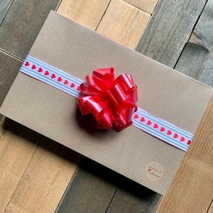 K&ouml;nnte beinhalten: Eine braune Geschenkbox mit einem wei&szlig;en Band und roten Herzen. Das Band ist zu einer gro&szlig;en roten Schleife gebunden. Die Box hat einen kleinen wei&szlig;en runden Aufkleber mit dem Text "handgefertigt mit Liebe".