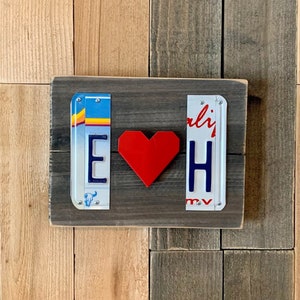 K&ouml;nnte beinhalten: Ein Holzschild mit zwei Nummernschildern, eines mit dem Buchstaben "E" und das andere mit dem Buchstaben "H". Ein rotes Herz befindet sich zwischen den Schildern. Das Schild befindet sich auf einem Holzgrund.