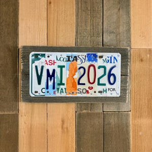 May include: A rectangular wall art piece featuring a license plate collage. The license plates are set in a gray wooden frame and mounted on a wooden background. The license plates display the letters "VMI" and the number "2026".