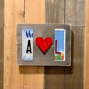 K&ouml;nnte beinhalten: Ein Holzschild mit zwei Nummernschildern, eines mit dem Buchstaben "A" und das andere mit dem Buchstaben "L". Ein rotes Herz ist zwischen den beiden Schildern platziert.
