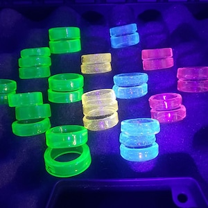 May include: A collection of colorful glass rings in various shades of green, blue, and pink. The rings are stacked and arranged on a dark surface, creating a vibrant and eye-catching display.