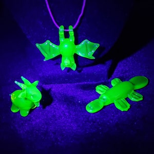 Op de afbeelding: Drie groene glazen dierenfiguren, een vleermuis, een vogelbekdier en een stier, onder een blacklight. De vleermuis hangt aan een koord.