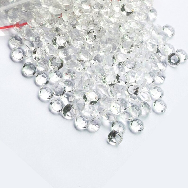 May include: A pile of clear, faceted, glass rhinestones. The rhinestones are scattered on a white surface.