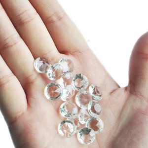 May include: A hand holding a pile of clear, faceted, round, glass gems. The gems are various sizes and are scattered on the palm of the hand.