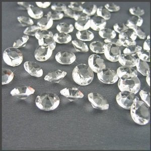 May include: A collection of numerous small, clear, faceted acrylic gems scattered across the frame. Each individual piece features a circular top with multiple triangular facets that catch and reflect light, creating a brilliant, diamond-like appearance.