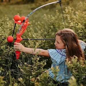 May include: A child aiming a bright orange toy bow and arrow in a field of green foliage. The bow has a colorful floral pattern and three orange balls. The child is wearing a blue and white striped shirt.