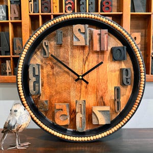 May include: A wooden clock with a black frame and a beaded edge. The clock face is made of wood and has numbers made from wooden blocks. The clock hands are black.