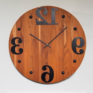 May include: A round wooden wall clock with a natural wood grain finish. The clock features large, black, stylized numbers and black metal hands. Small black dots mark the hours. The clock is approximately 12 inches in diameter.