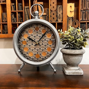 May include: A silver clock with a face made of coins. The clock has three legs and is sitting on a wooden table. A small potted plant is to the right of the clock.