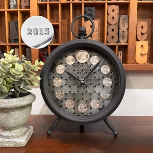 May include: Black metal clock with a round face filled with pennies. The clock has a loop at the top and three legs. The hands are black. A small, circular metal piece with the number "2015" is in the upper left corner.