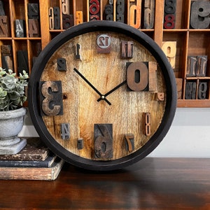 May include: A round wooden clock with black hands and a black frame. The clock face is made of wood and has numbers made of wood blocks. The clock is hanging on a wall with a wooden shelf behind it.