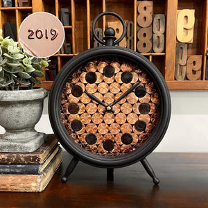 May include: Black metal clock with a copper penny face and black hands. The clock has a circular shape with a small handle at the top and three legs. The year "2019" is stamped on a circular tag.