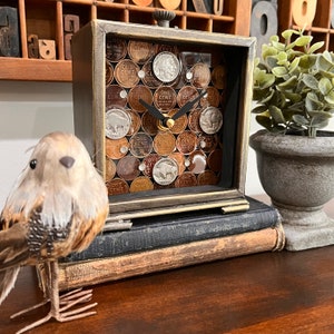 May include: A square clock with a wood frame and glass face. The clock face is made of a mosaic of vintage US one-cent coins. The clock is sitting on a stack of books.