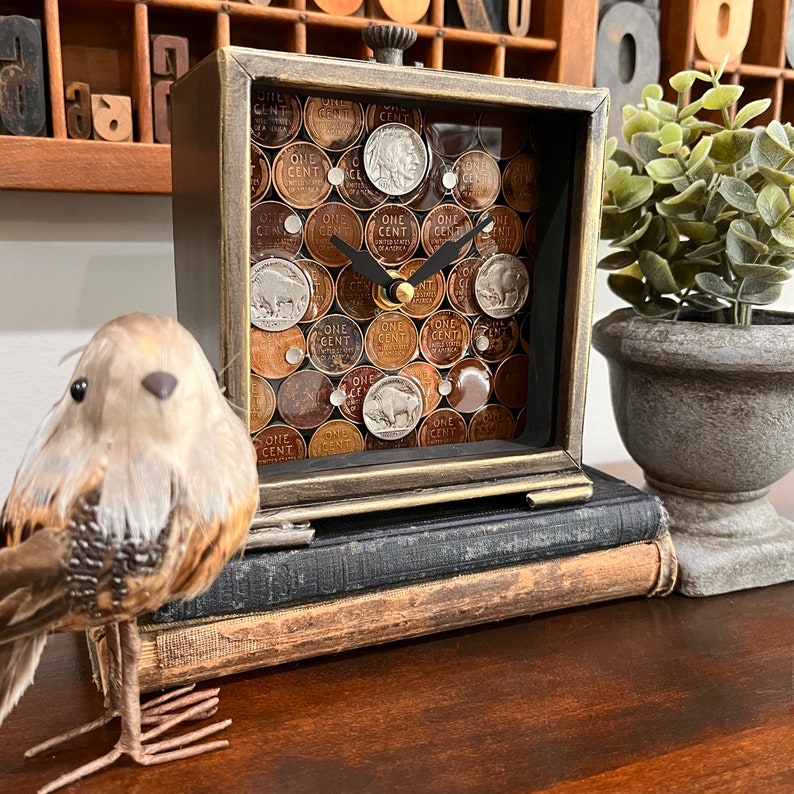 May include: A square clock with a wood frame and glass face. The clock face is made of a mosaic of vintage US one-cent coins. The clock is sitting on a stack of books.