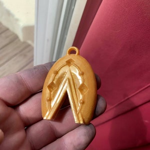 Puede incluir: Un colgante de color dorado con un diseño geométrico. El colgante tiene forma de lágrima y tiene un pequeño bucle en la parte superior para sujetar una cadena.