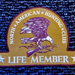 Könnte beinhalten: Ein goldfarbener Aufnäher mit der Aufschrift "NORTH AMERICAN HUNTING CLUB" und "LIFE MEMBER". Der Aufnäher zeigt einen Adler mit Gewehr und Bogen. Text und Adler sind in Burgunderrot und Weiß gehalten.